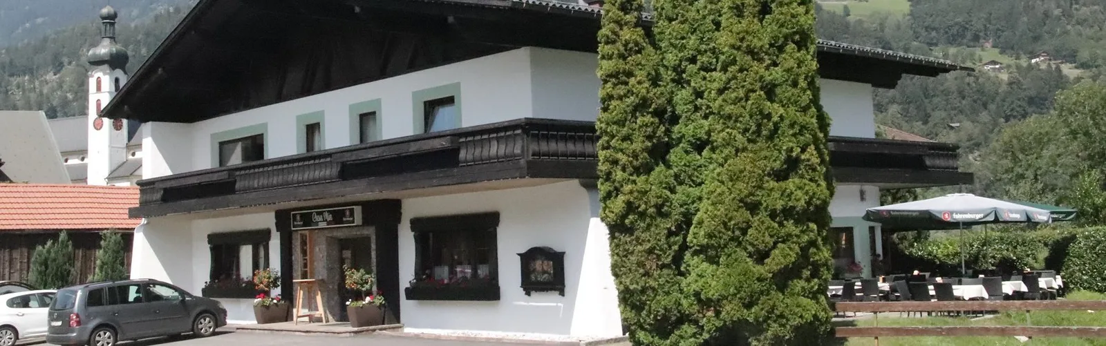 Das Haus der Pizzeria Casa Mia von der Straße, mit Schild über dem Eingang, Gastgarten und Kirchturm dahinter