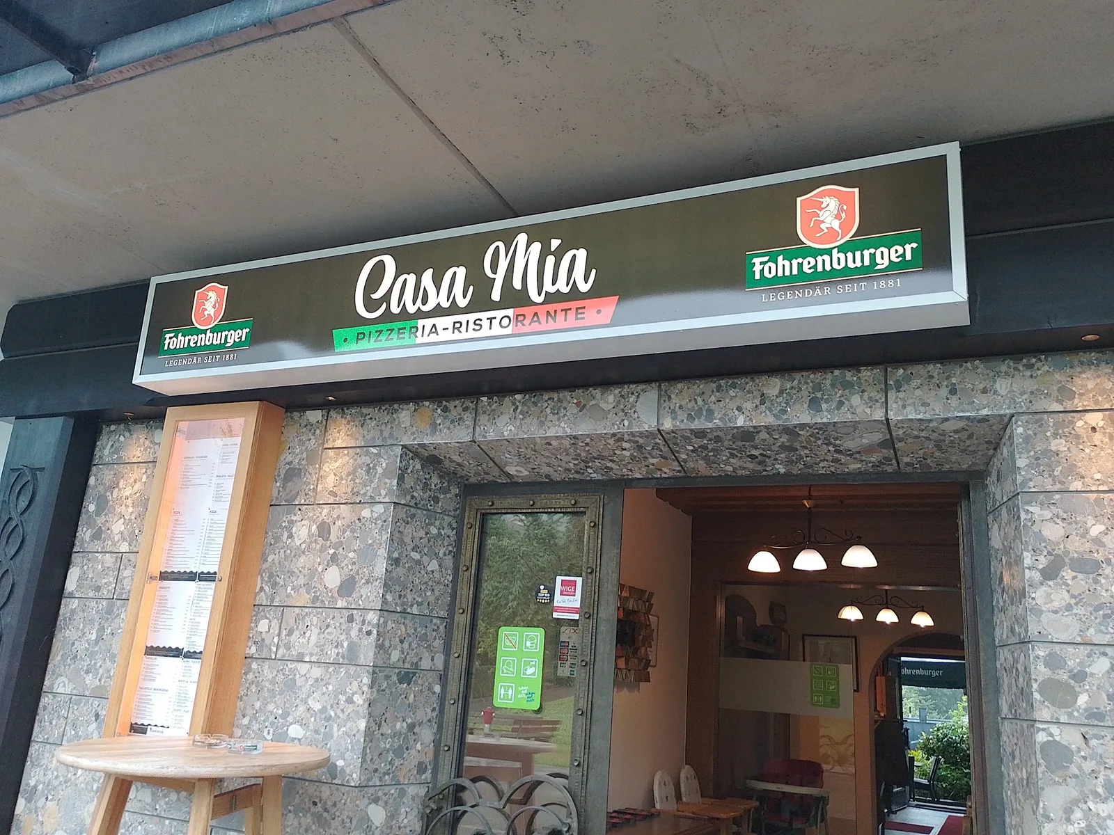 Beleuchtetes Schild „Casa Mia Pizzeria-Ristorante“ über dem Eingang, daneben der Schaukasten mit der Speisekarte